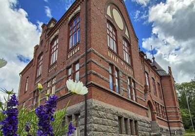 Lahden yhteiskoulun punatiilinen 1899 rakennuttu goottilaistyylinen koulurakennus nimeltä Museo. Taustalla sininen taivas ja valkoisia pilviä. Etualalla kesäkukkia.
